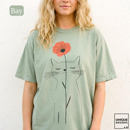 May include: A sage green short-sleeve t-shirt featuring a black line art illustration of a cat with closed eyes. A single orange poppy flower is positioned above the cat's head, with its stem extending downward along the cat's body.