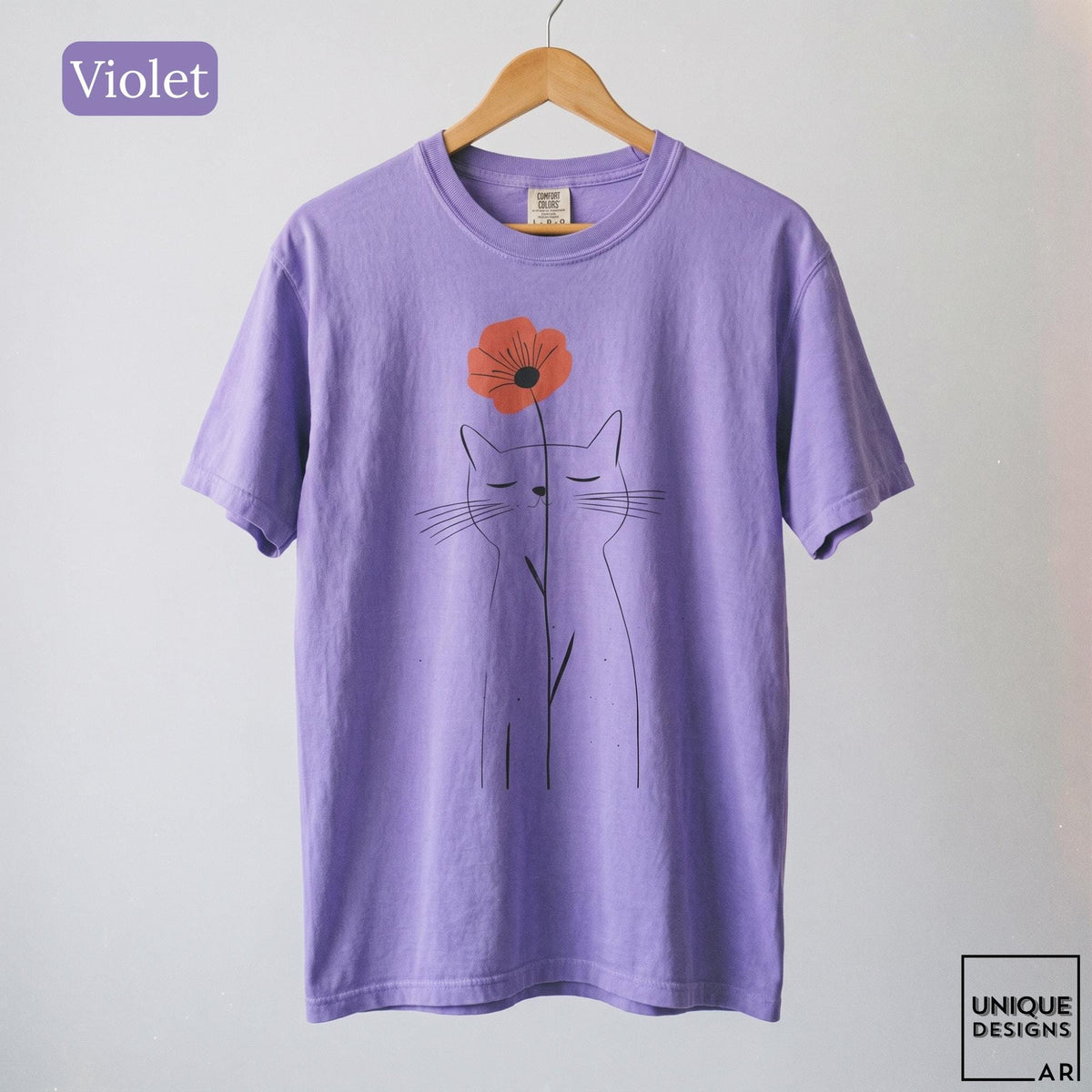May include: A violet short-sleeve t-shirt featuring a black line art illustration of a cat with closed eyes. A single orange flower is positioned above the cat's head. The text "Violet" appears in the top left corner, and "UNIQUE DESIGNS ARI" is in the bottom right.