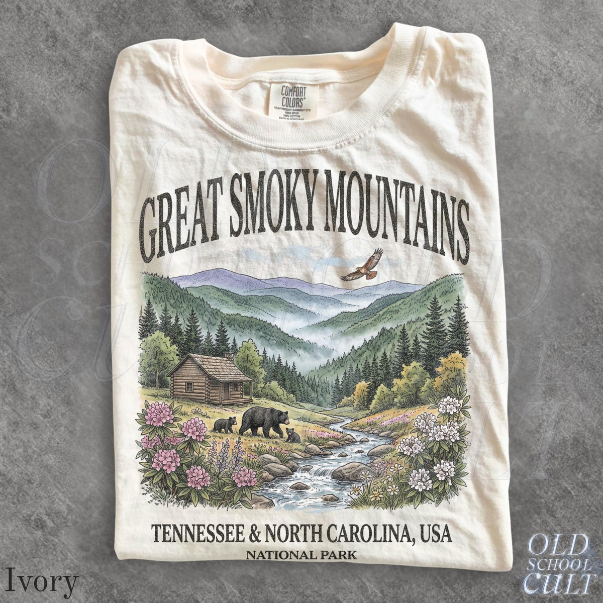 May include: An ivory t-shirt featuring a graphic print of the Great Smoky Mountains. The design includes a log cabin, a stream, black bears, and a flying eagle amidst pine trees and flowers, with the text Great Smoky Mountains and Tennessee & North Carolina, USA National Park.