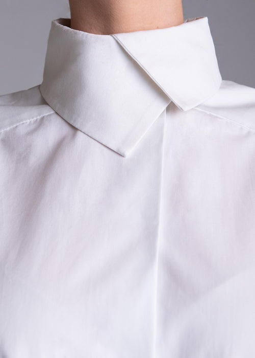 May include: A crisp white button-down shirt featuring a structured high collar with a pointed fold. The garment is made of a smooth, opaque fabric with a clean, minimalist design, showcasing a concealed placket and sharp, tailored shoulder seams.