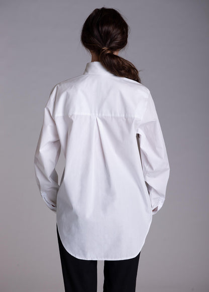 May include: A crisp white long-sleeved button-down shirt featuring a classic collar, a central back box pleat, and a curved hemline. The shirt is worn over plain black trousers, showcasing a clean, structured silhouette with long sleeves.