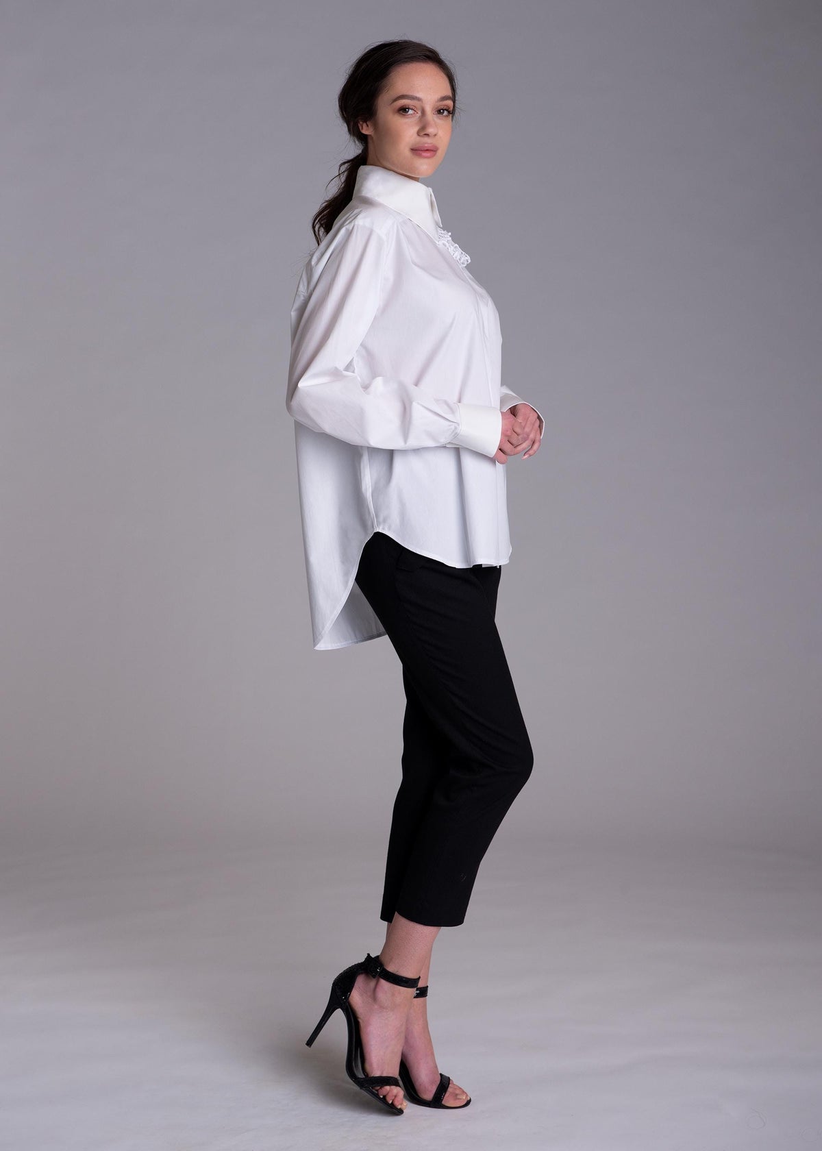 May include: A crisp white long-sleeved button-down shirt with a structured collar and cuffs, paired with slim-fit black trousers that end above the ankle. The shirt features a high-low hemline and a subtle ruffled detail along the front placket.