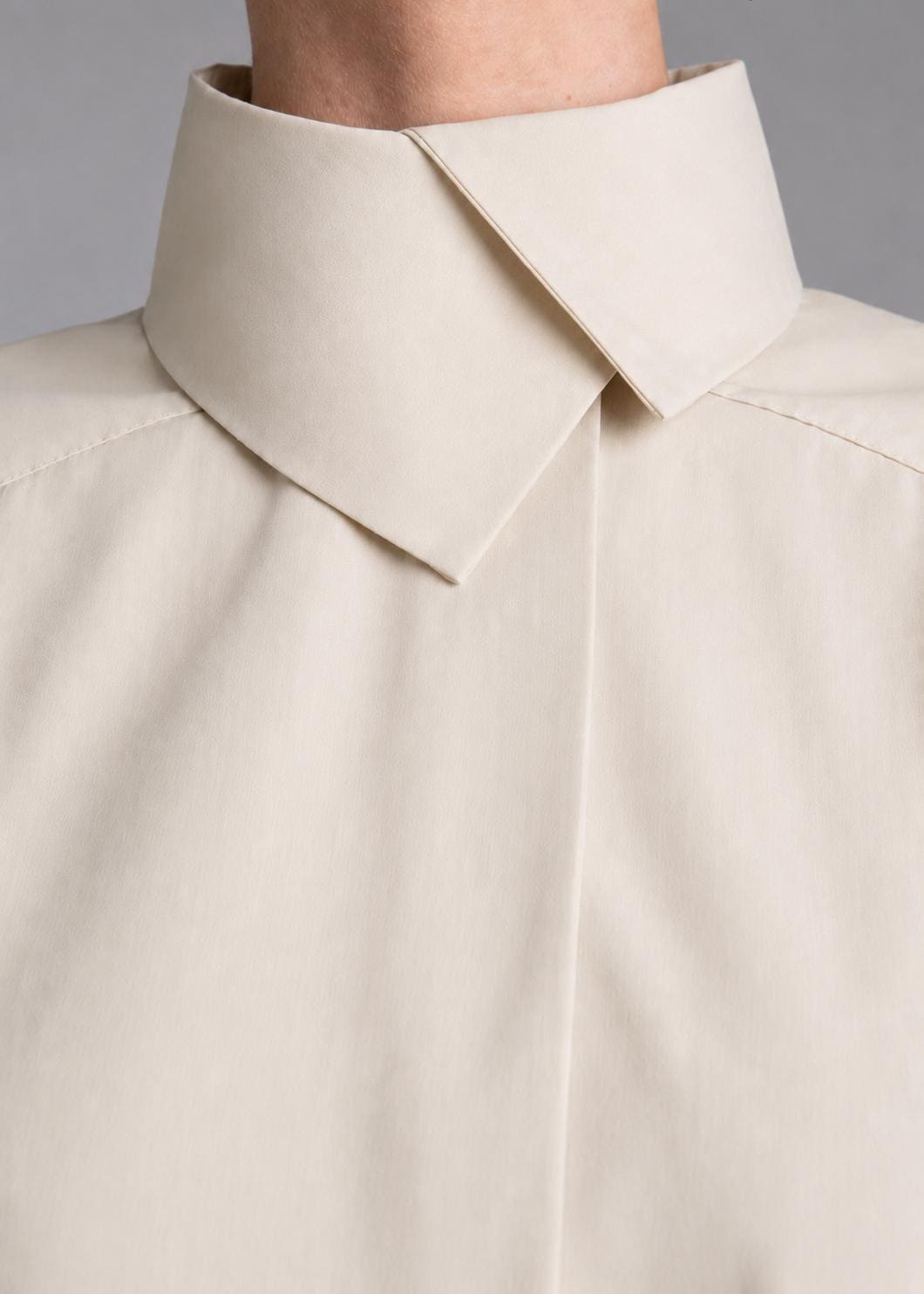 May include: A light beige button-down shirt featuring a high, structured collar with a sharp, pointed fold. The garment has a clean, smooth texture and a concealed placket design, focusing on the neckline and upper chest area of the shirt.