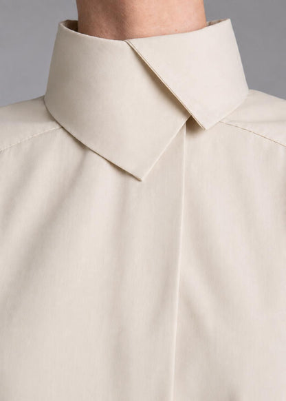 May include: A light beige button-down shirt featuring a high, structured collar with a sharp, pointed fold. The garment has a clean, smooth texture and a concealed placket design, focusing on the neckline and upper chest area of the shirt.