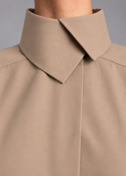 May include: A beige collared shirt featuring a high, structured stand-up collar with a pointed fold-over detail. The garment has a smooth, matte finish and a concealed front placket, showcasing clean lines and a minimalist construction throughout.