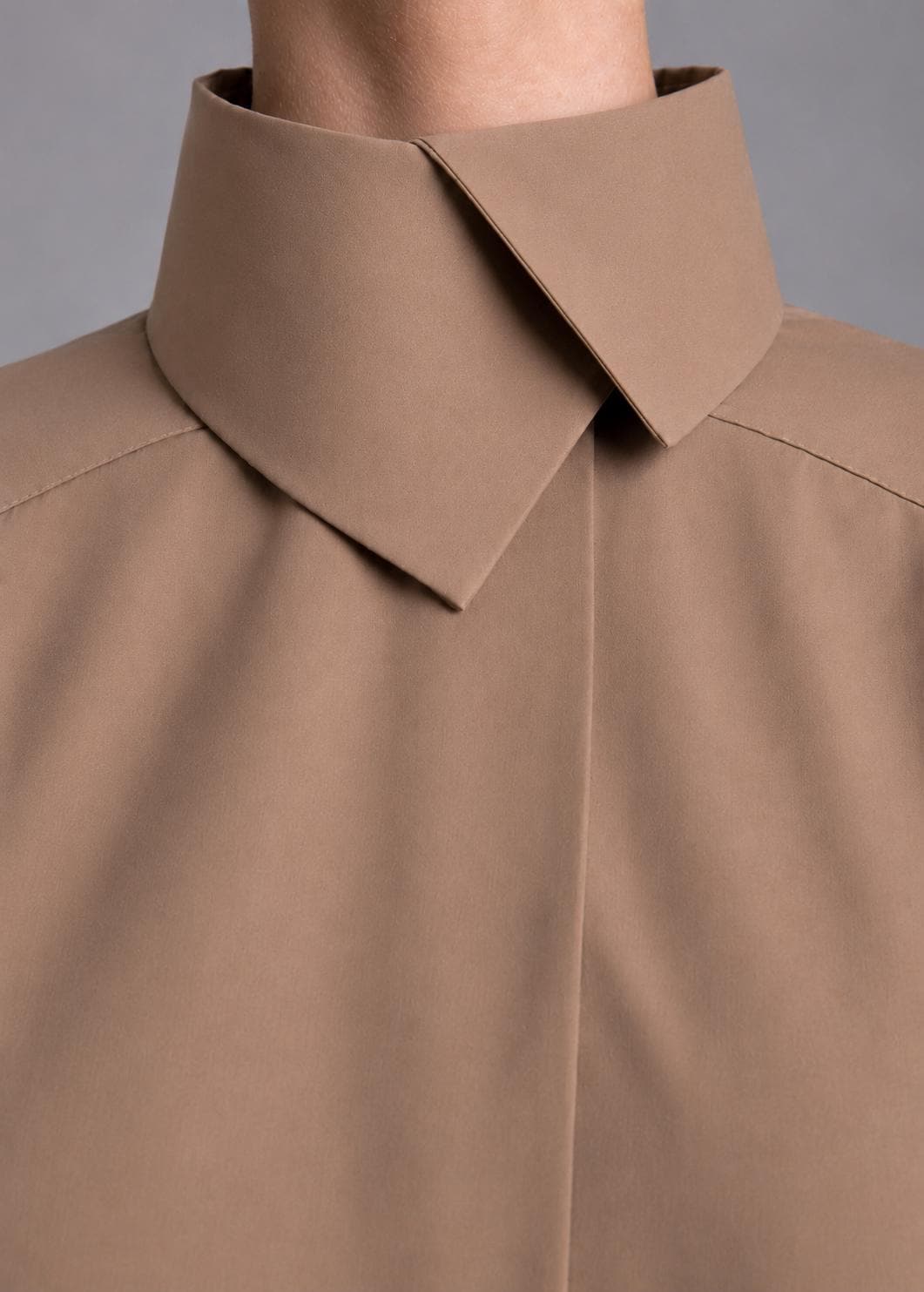 May include: A solid tan shirt featuring a high, structured collar with a sharp, overlapping point design. The garment has a smooth, matte finish and clean, minimalist lines, focusing on the tailored construction of the neckline and shoulder seams.