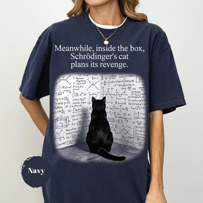 May include: A navy blue t-shirt featuring a graphic of a black cat sitting in front of a wall covered in white physics equations and diagrams. The text above the cat reads, "Meanwhile, inside the box, Schrödinger's cat plans its revenge."