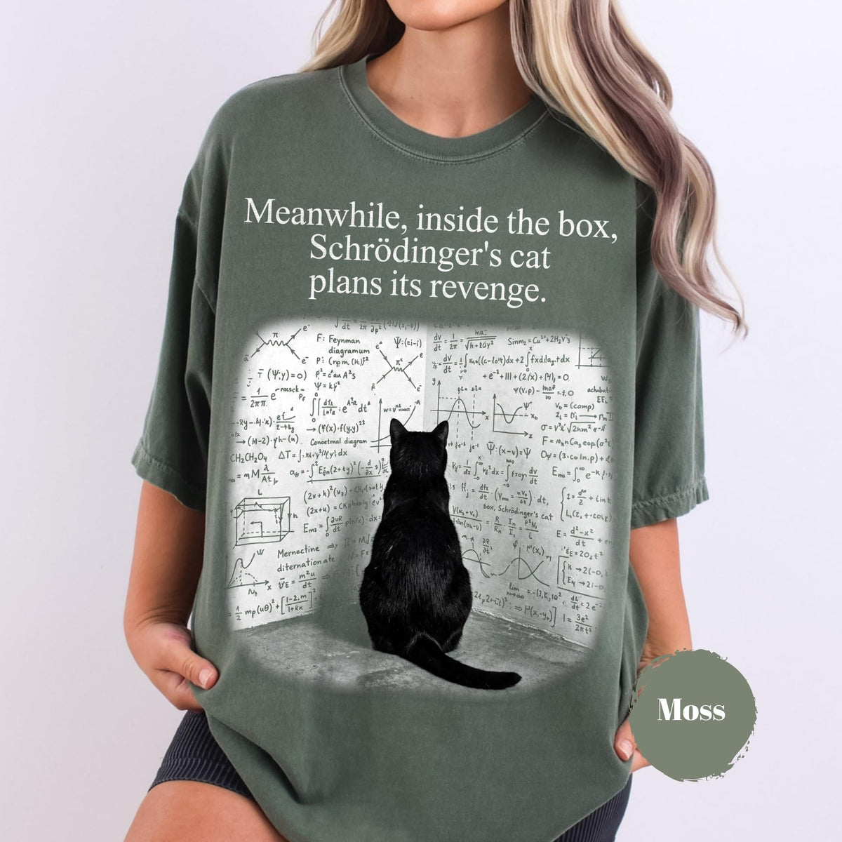 May include: A dark olive green t-shirt featuring a graphic of a black cat sitting in front of a wall covered in complex physics equations and diagrams. The text above the graphic reads, "Meanwhile, inside the box, Schrödinger's cat plans its revenge."