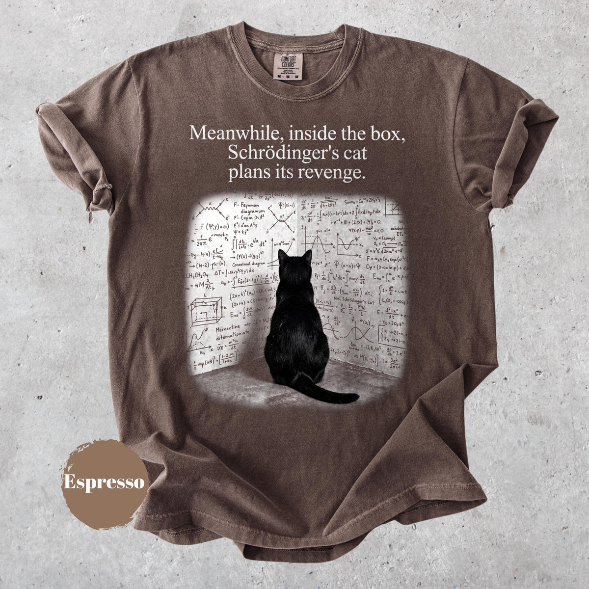 May include: A brown short-sleeved t-shirt featuring a graphic of a black cat sitting before a wall covered in complex physics equations. The text above the graphic reads, "Meanwhile, inside the box, Schrödinger's cat plans its revenge." A circular brown tag labeled "Espresso" is visible.