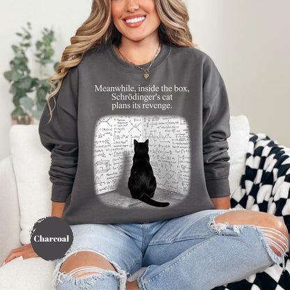 May include: A charcoal gray crewneck sweatshirt featuring a graphic of a black cat sitting before a wall covered in complex mathematical equations. The text above the cat reads, "Meanwhile, inside the box, Schrödinger's cat plans its revenge."