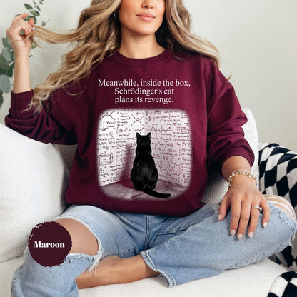 May include: A maroon long-sleeve sweatshirt featuring a graphic of a black cat sitting before a wall covered in complex mathematical equations. The text above the graphic reads, "Meanwhile, inside the box, Schrödinger's cat plans its revenge."