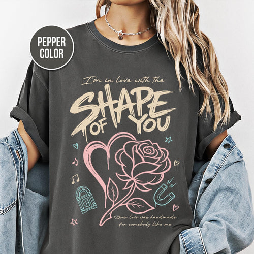 Shape of You” text with heart and rose illustration, music icons and pastel accents printed on a pepper Comfort Colors oversized T-shirt.