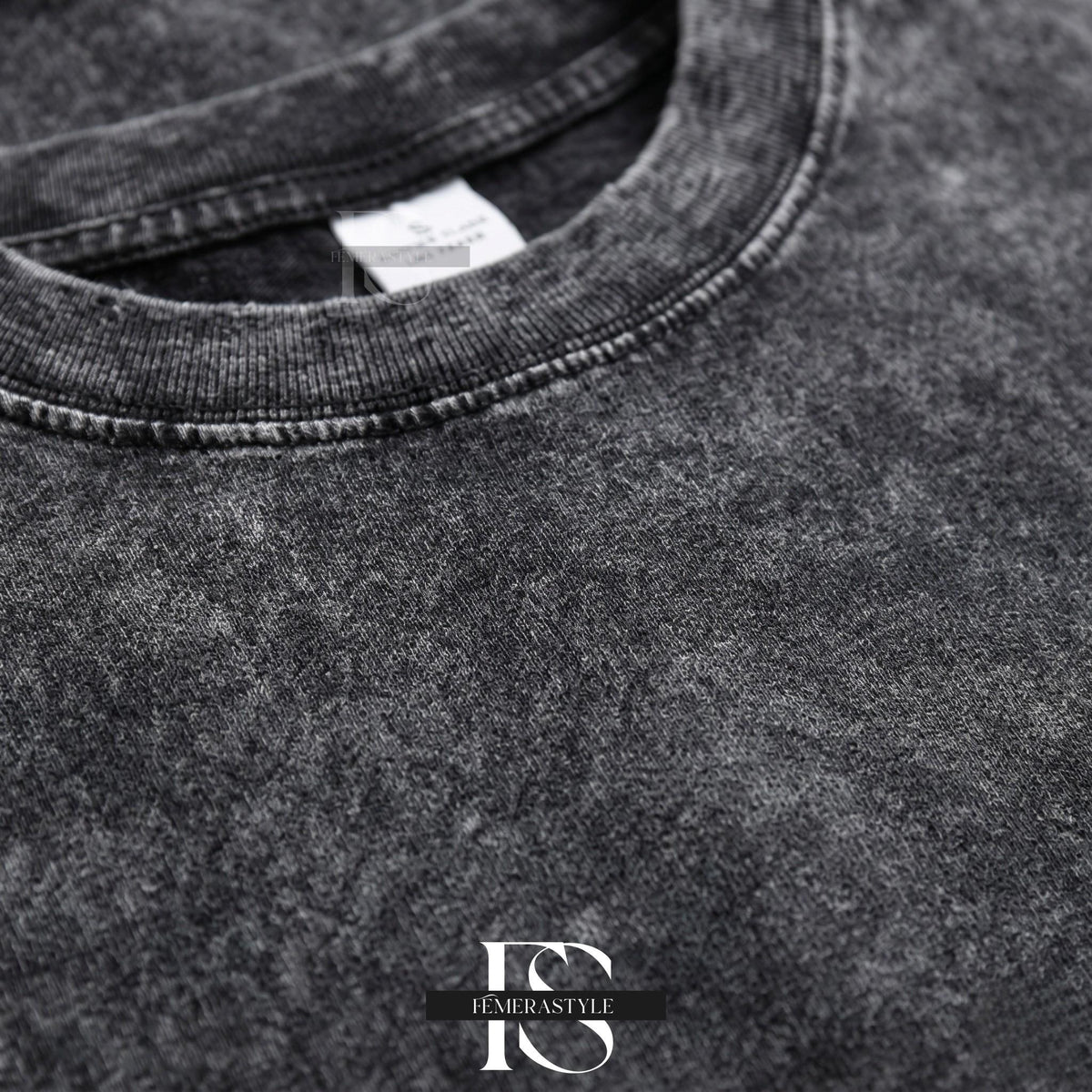 May include: Close-up of a charcoal gray t-shirt with a textured, acid wash effect. The crew neck and seams are visible, with a small white tag near the collar. The bottom right corner features the text "IEMERASTYLE" in a stylized font.