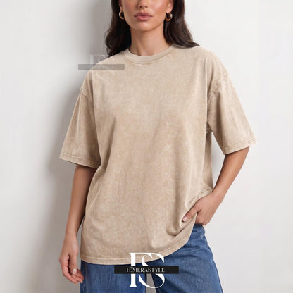 May include: A beige, oversized t-shirt with a crew neck and short sleeves. The shirt has a faded, textured appearance. The model is wearing blue jeans. The shirt is from the brand "IEMERASTYLE".