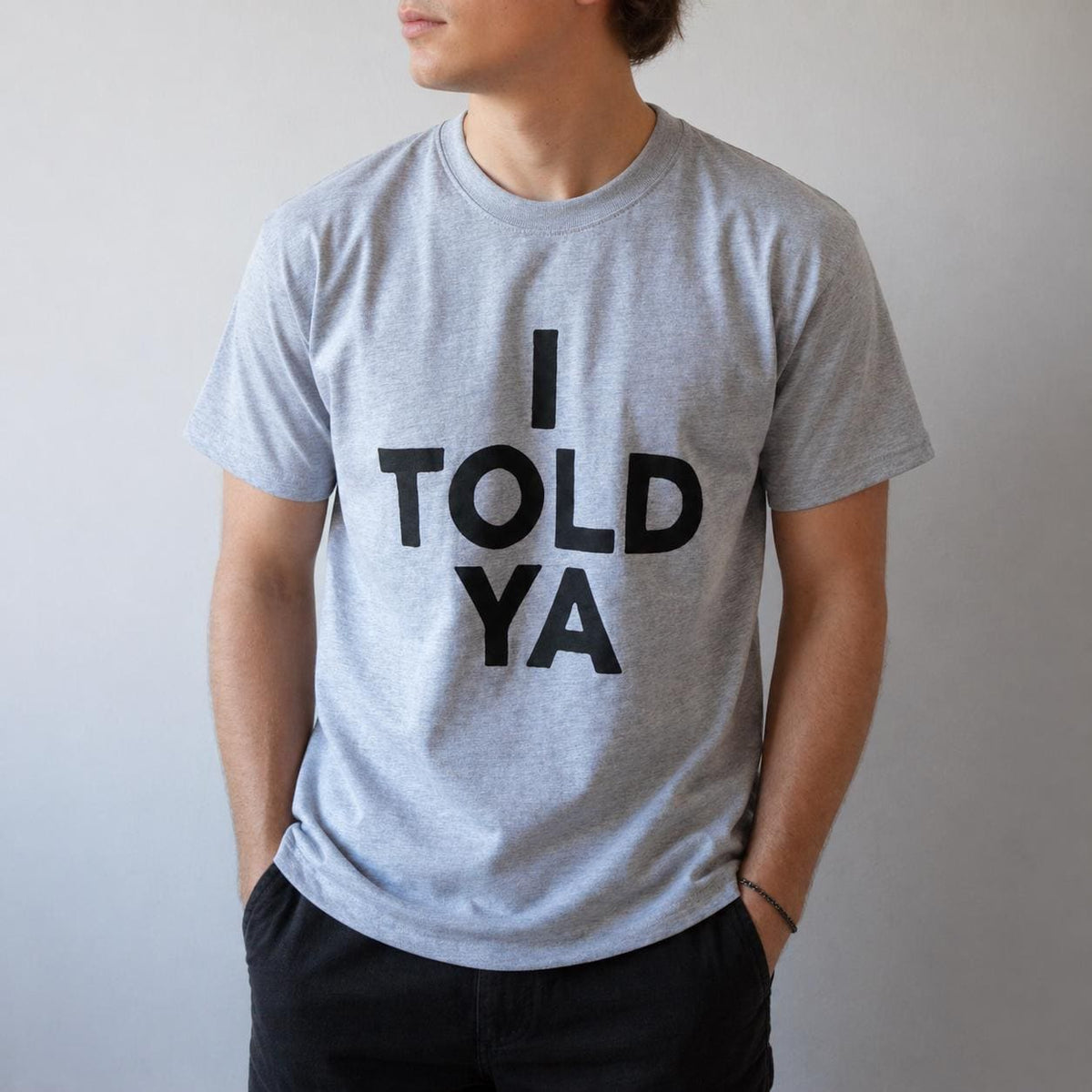 May include: A light gray t-shirt with the text "I TOLD YA" in bold black letters. The shirt has short sleeves and a crew neck. The person is wearing black pants.