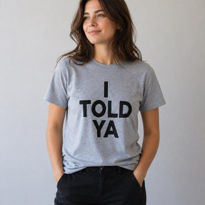 May include: A gray t-shirt with the text "I TOLD YA" in bold black letters. The shirt is short-sleeved and appears to be made of a soft material. The person is wearing black pants.