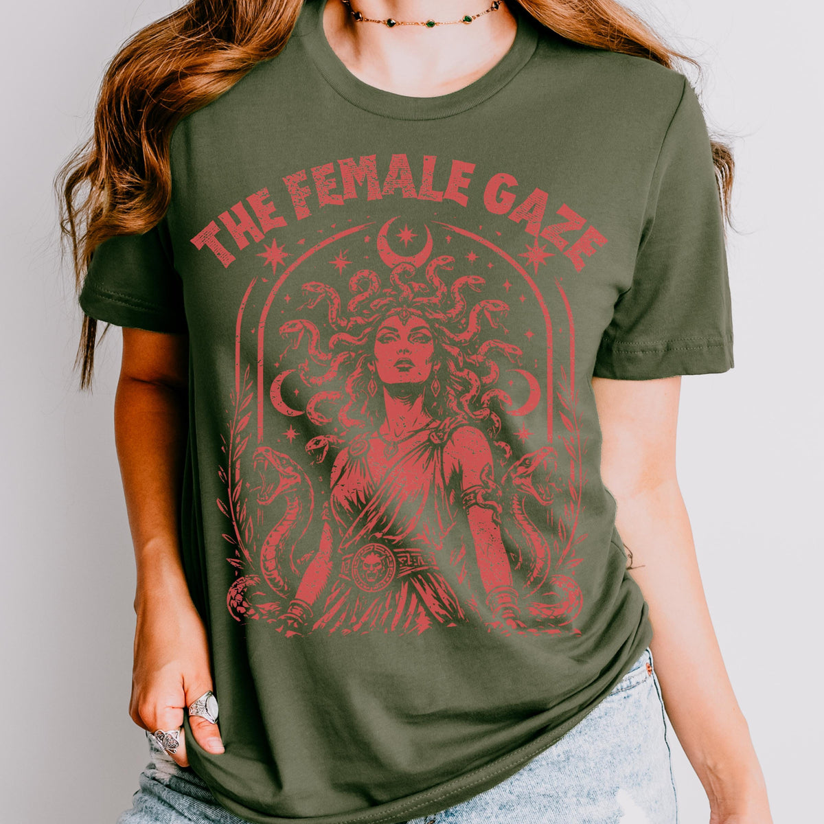 May include: Olive green t-shirt with a red graphic of Medusa and the text "THE FEMALE GAZE". The graphic features Medusa with snakes for hair, surrounded by snakes and crescent moons. The design is a vintage style.