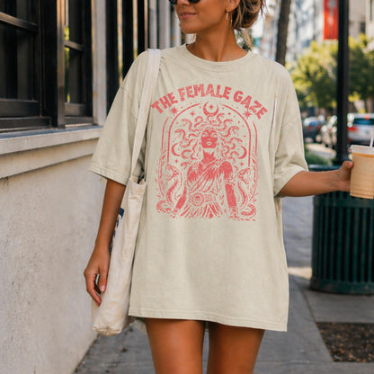 May include: Cream-colored oversized t-shirt with a red graphic of Medusa and the text "THE FEMALE GAZE". The graphic features a woman with snakes for hair, surrounded by decorative elements. The shirt is a casual, comfortable style.