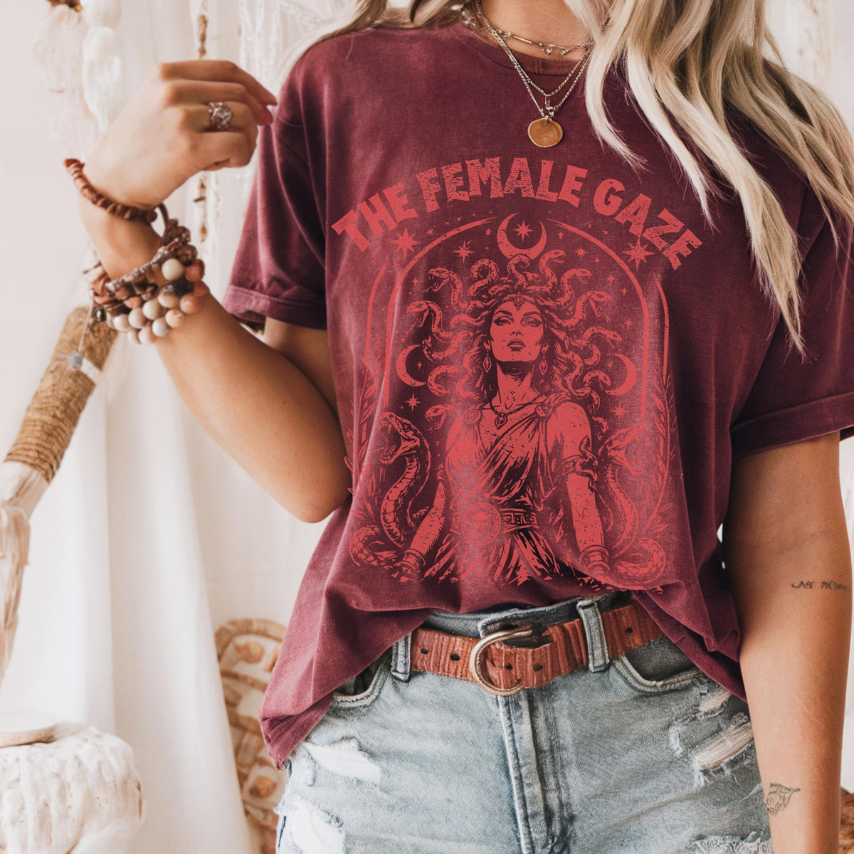 May include: Burgundy t-shirt with a graphic of Medusa and the text "THE FEMALE GAZE" in red. The design includes snakes, stars, and a crescent moon. The shirt is paired with denim shorts and a brown belt.