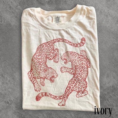 May include: Ivory-colored t-shirt featuring a red illustration of two leopards in a dynamic pose. The shirt has a crew neck and short sleeves. The brand name "Comfort Colors" is visible on the tag. The word "ivory" is printed at the bottom right.