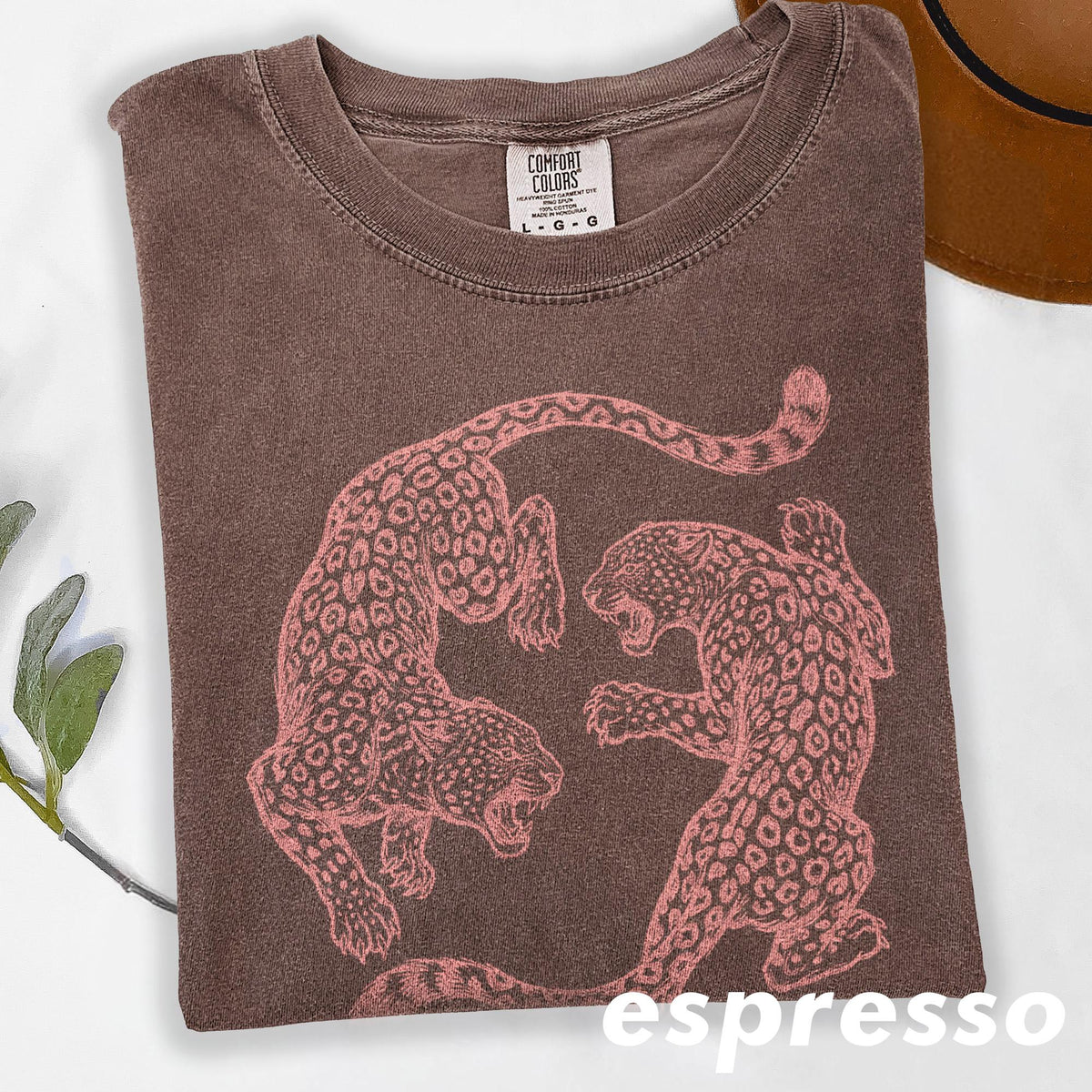 May include: Espresso-colored t-shirt featuring a pink leopard design. The shirt has a crew neck and short sleeves. The graphic shows two leopards facing each other. The shirt is folded and has a tag that says "Comfort Colors".