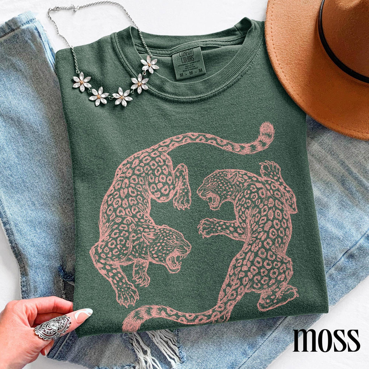 May include: Moss green sweatshirt featuring a pink leopard print design. The sweatshirt has a crew neck and long sleeves. A silver necklace with flower-shaped pendants is draped on the shirt. The word "moss" is printed on the bottom right.
