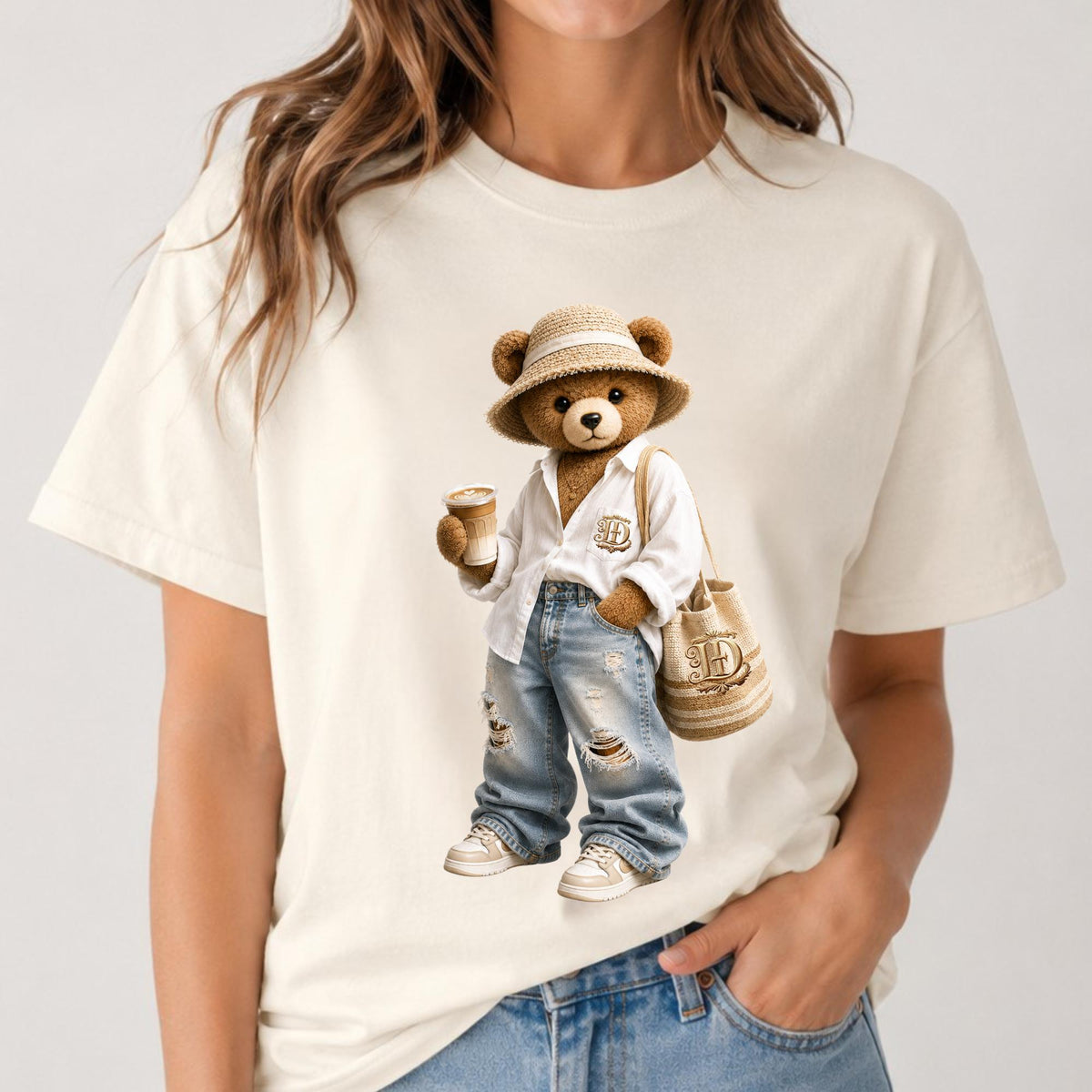 May include: Cream-colored t-shirt featuring a cartoon bear wearing a straw hat, white shirt, ripped jeans, and sneakers. The bear holds a coffee cup and a tote bag with a logo. The bear is a stylish character.