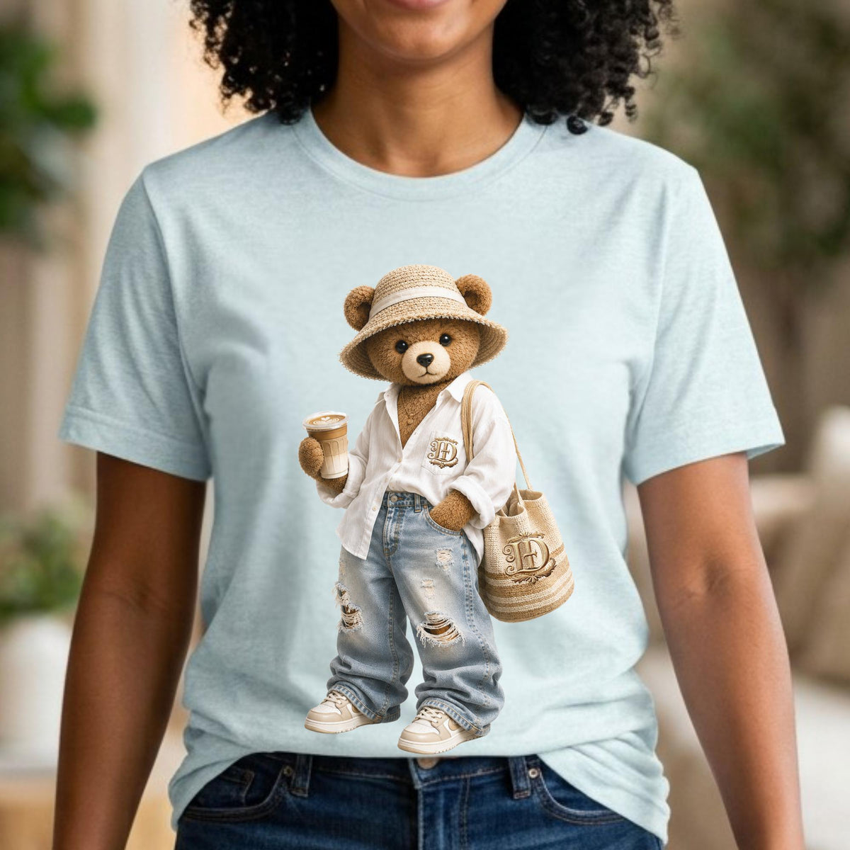 May include: Light blue t-shirt featuring a cartoon bear wearing a white shirt, ripped jeans, and a straw hat. The bear is holding a coffee cup and a tote bag with the letter "H" on it. The bear is wearing white sneakers.