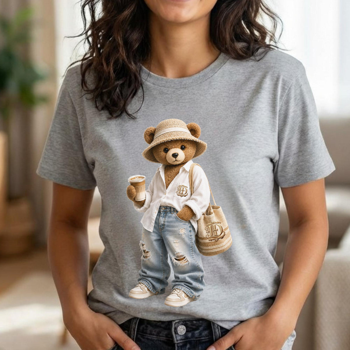 May include: A gray t-shirt featuring a cartoon bear wearing a straw hat, white shirt, ripped jeans, and sneakers. The bear is holding a coffee cup and a beige tote bag with a monogram. The bear is a light brown color.