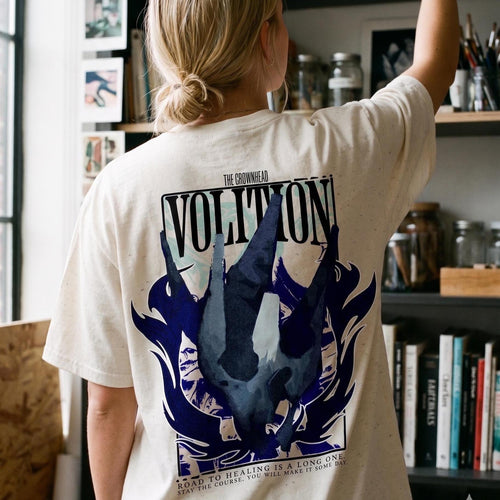 May include: An off-white short-sleeve t-shirt featuring a large graphic on the back. The design includes the word VOLITION in bold black letters, a dark blue abstract shape, and the text: THE CROWNHEAD ROAD TO HEALING IS A LONG ONE.