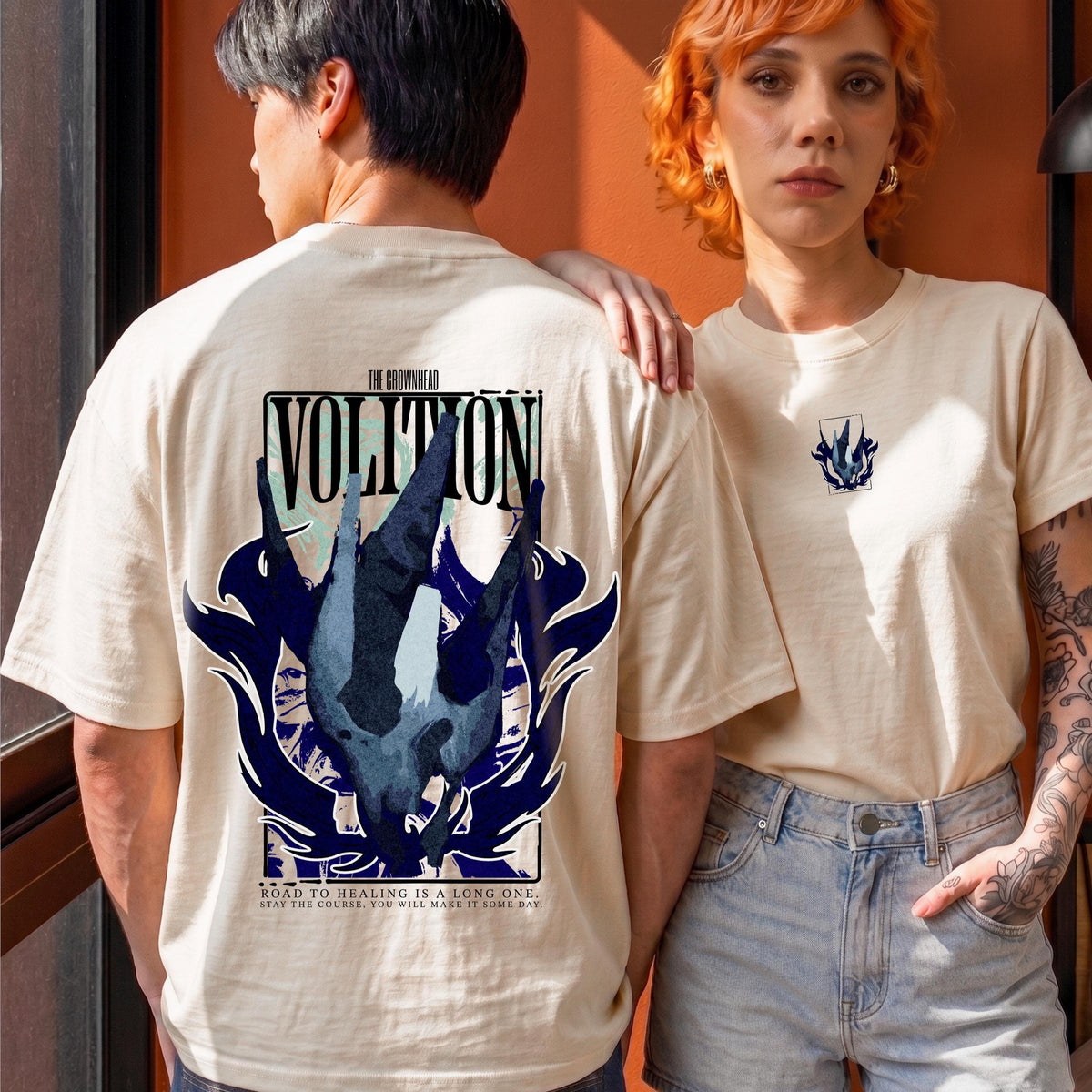 May include: A beige short-sleeve t-shirt featuring a large graphic on the back with the text VOLITION and THE GROWNHEAD. The design includes a dark blue abstract shape and the text ROAD TO HEALING IS A LONG ONE. STAY THE COURSE. YOU WILL MAKE IT SOME DAY.