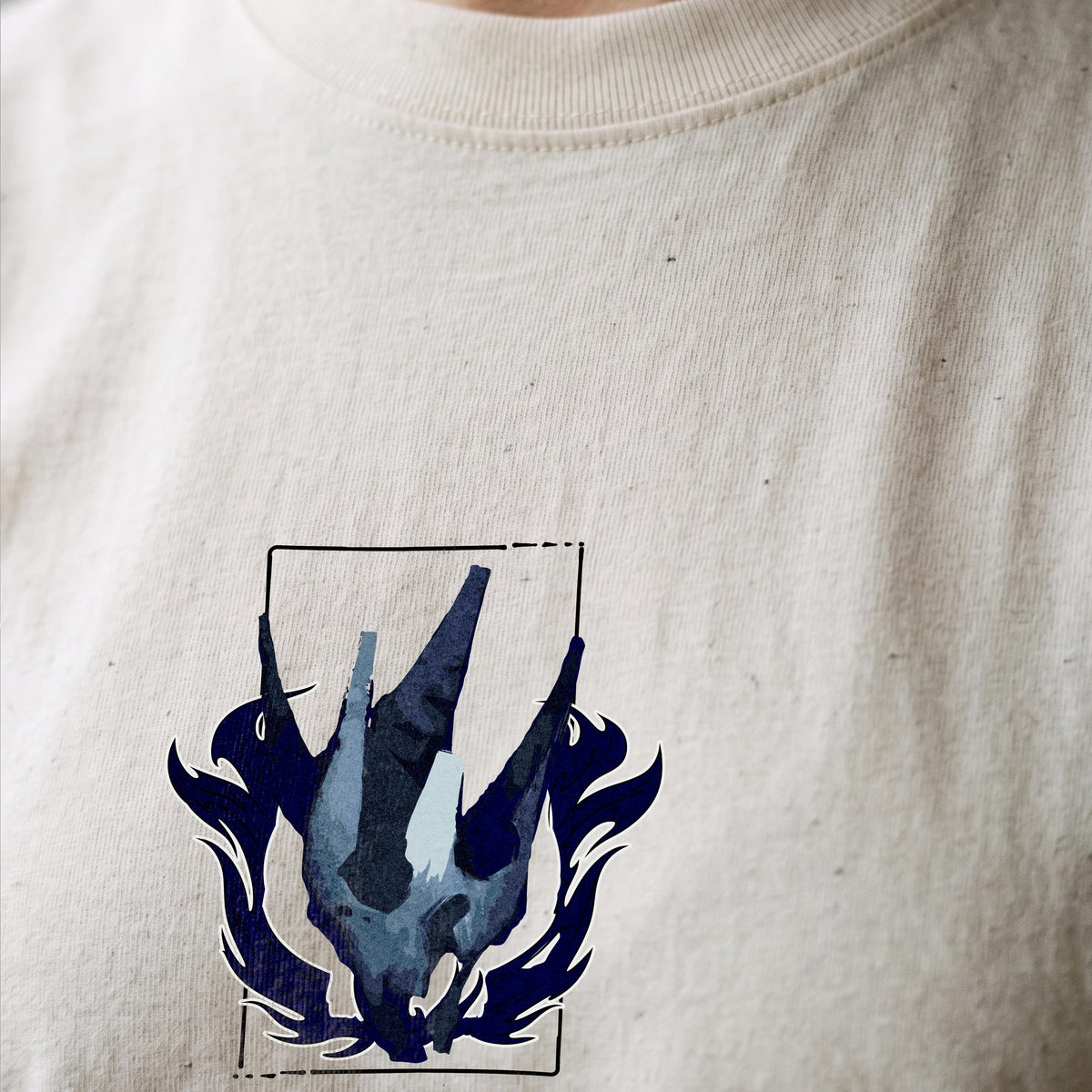 May include: A cream-colored t-shirt featuring a graphic print of a dark blue, stylized, angular, abstract shape resembling a bird or wing structure, enclosed within a thin black rectangular frame with decorative flame-like accents at the base.