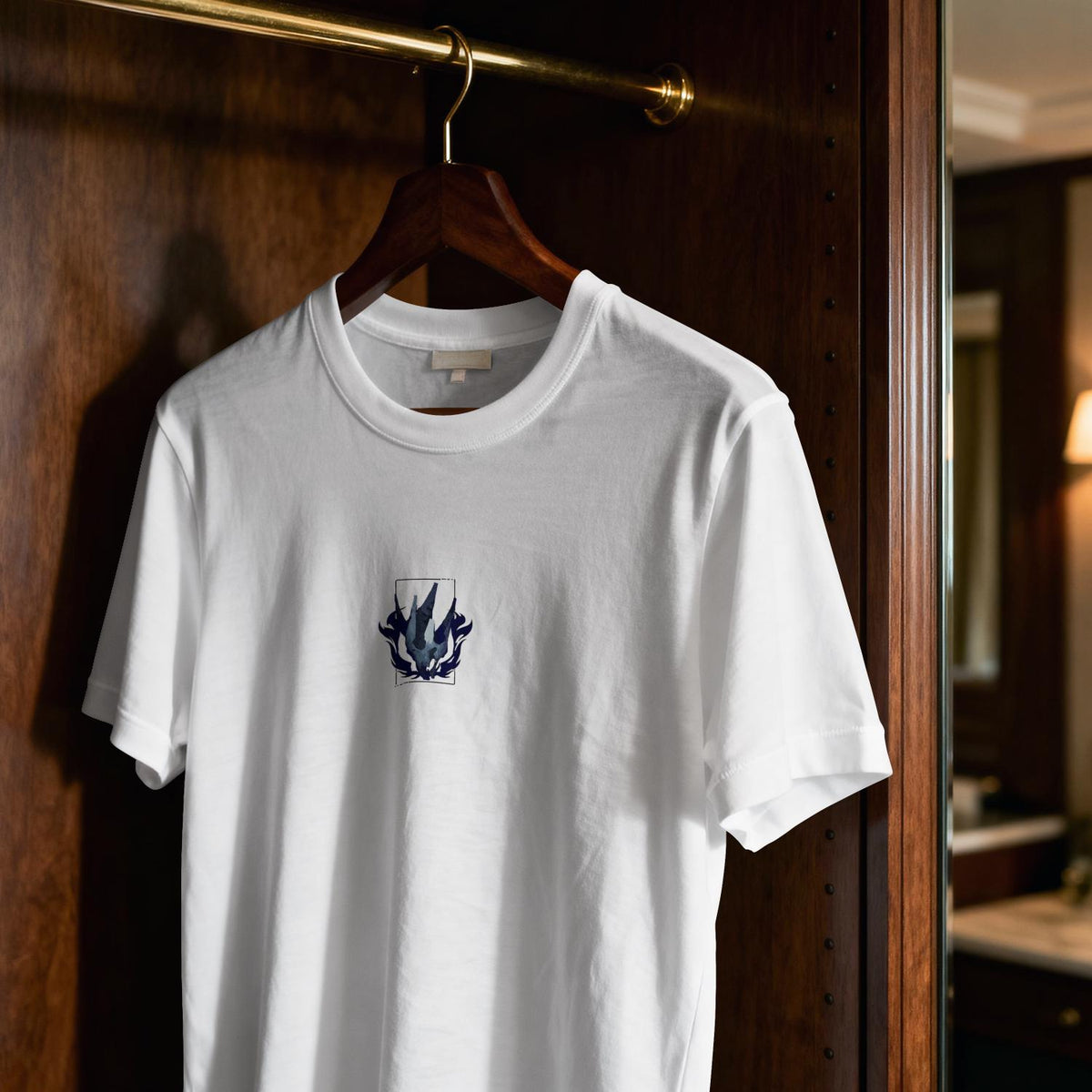 May include: A white short-sleeved crew neck t-shirt featuring a small, centered graphic on the chest. The design consists of a dark blue and grey abstract, flame-like shape enclosed within a thin rectangular border, printed on the fabric.