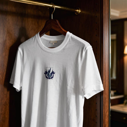 May include: A white short-sleeved crew neck t-shirt featuring a small, centered graphic on the chest. The design consists of a dark blue and grey abstract, flame-like shape enclosed within a thin rectangular border, printed on the fabric.