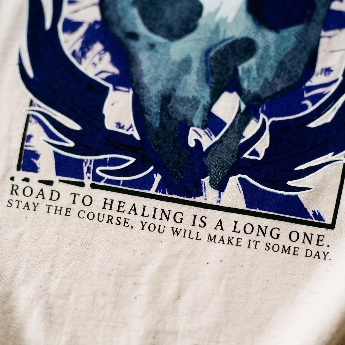 May include: A cream-colored fabric featuring a dark blue graphic print of a stylized bird or creature with wings. Below the graphic, black text reads: ROAD TO HEALING IS A LONG ONE. STAY THE COURSE, YOU WILL MAKE IT SOME DAY. The design is framed.