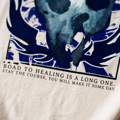 May include: A cream-colored fabric featuring a dark blue graphic print of a stylized bird or creature with wings. Below the graphic, black text reads: ROAD TO HEALING IS A LONG ONE. STAY THE COURSE, YOU WILL MAKE IT SOME DAY. The design is framed.