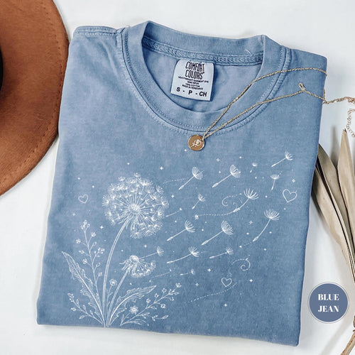May include: Blue jean-colored t-shirt with a white dandelion graphic. The dandelion is blowing seeds, with hearts and stars scattered around. The shirt has a Comfort Colors tag and a gold necklace.