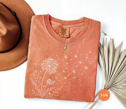 May include: Coral-colored t-shirt with a white dandelion graphic. The design features a detailed dandelion with seeds blowing away, accompanied by small hearts. A gold necklace with a pendant rests on the shirt. A brown hat and dried palm leaves are also in the image.