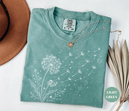 May include: Light green t-shirt with a dandelion graphic. The white design shows a dandelion with seeds blowing away, along with hearts and small flowers. A silver necklace with a pendant rests on the shirt. The shirt is size S-P-CH.