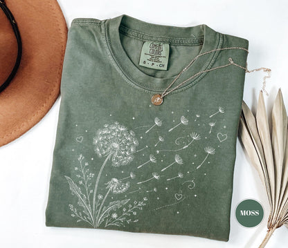 May include: Moss green t-shirt with a dandelion graphic. The design features a detailed white illustration of a dandelion with seeds blowing away, accompanied by hearts and stars. The shirt has a crew neck and short sleeves.