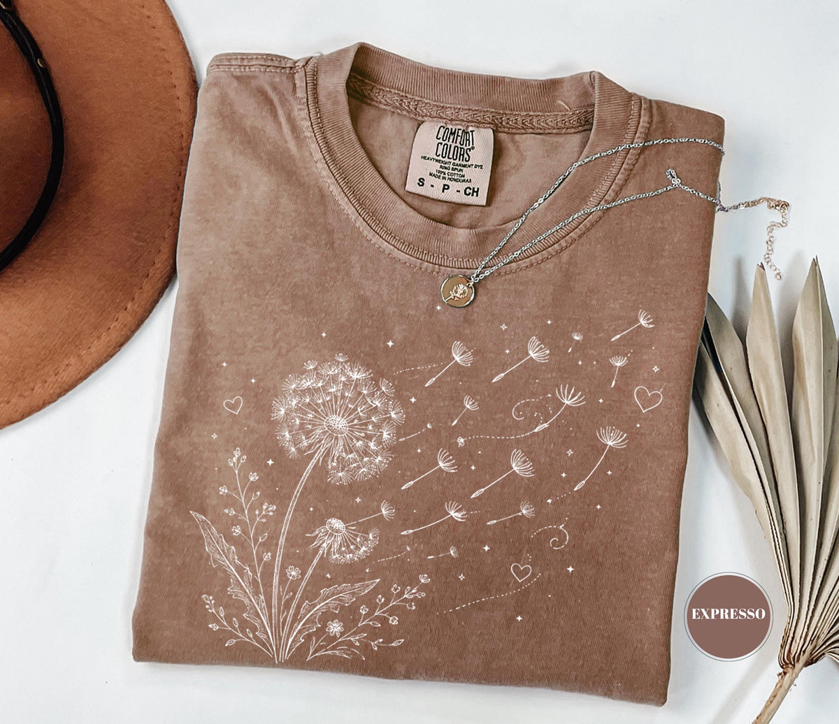 May include: A brown Comfort Colors t-shirt featuring a white dandelion design with seeds blowing in the wind. A silver necklace with a small pendant rests on the shirt. The shirt has a crew neck and short sleeves.