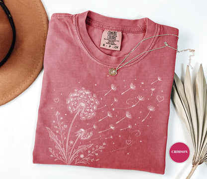 May include: Crimson-colored t-shirt featuring a dandelion design with seeds blowing in the wind. The shirt has a crew neck and short sleeves. A silver necklace with a small pendant rests on the shirt.