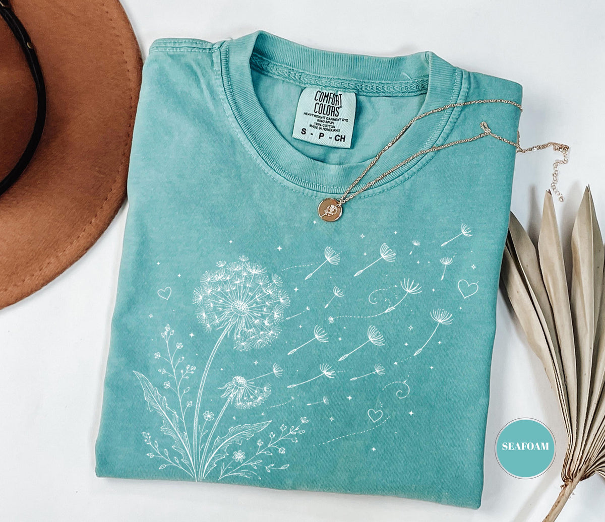 May include: Seafoam green t-shirt with a dandelion design. The white graphic shows a dandelion with seeds blowing away, along with hearts and small flowers. A gold necklace with a pendant rests on the shirt. The tag reads "Comfort Colors".