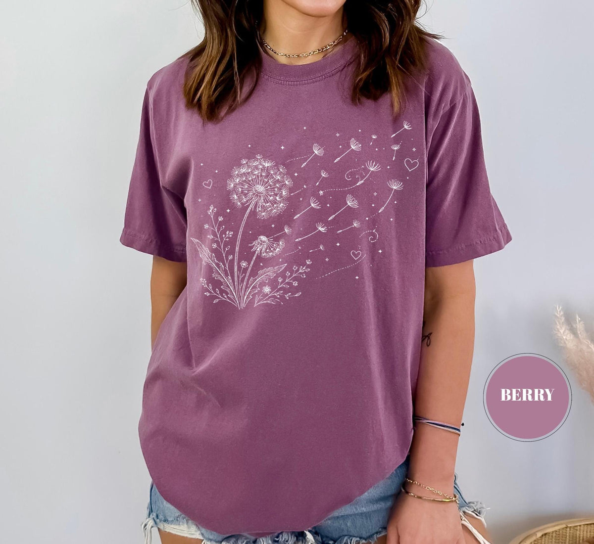 May include: Berry-colored t-shirt featuring a white dandelion design. The graphic shows a detailed dandelion with seeds blowing away, accompanied by small hearts. The shirt has short sleeves and a relaxed fit.