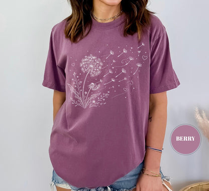 May include: Berry-colored t-shirt featuring a white dandelion design. The graphic shows a detailed dandelion with seeds blowing away, accompanied by small hearts. The shirt has short sleeves and a relaxed fit.