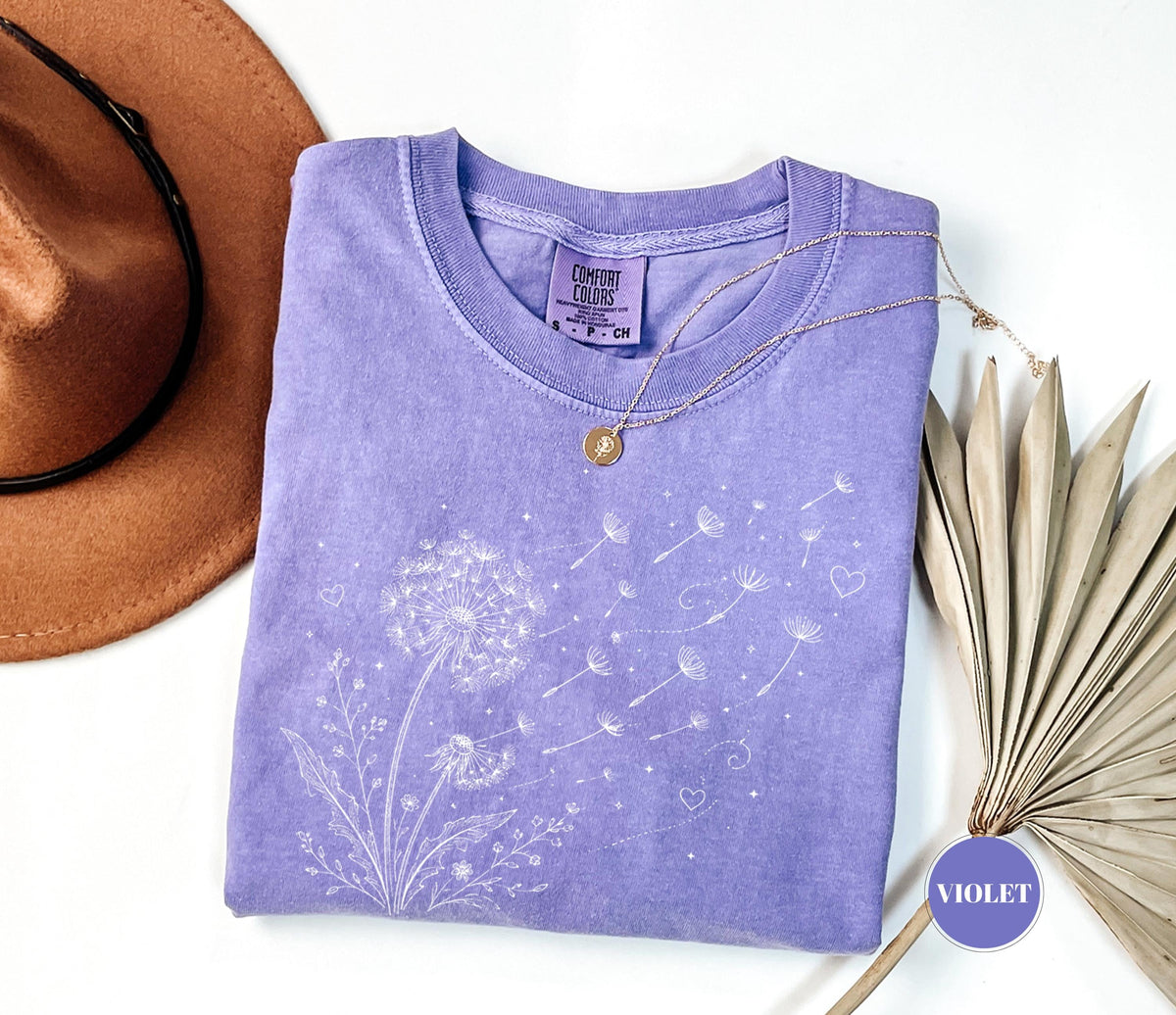 May include: A violet Comfort Colors t-shirt featuring a white dandelion design with seeds blowing away. A gold necklace and a brown hat are next to the shirt. The shirt has a crew neck and a small tag.
