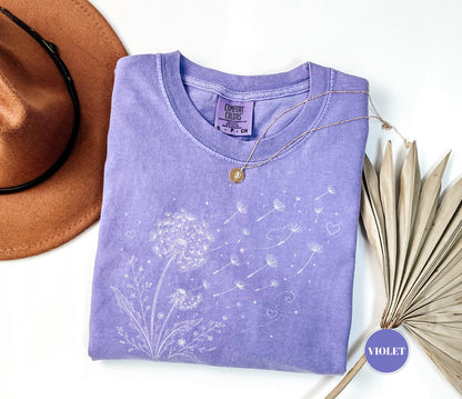 May include: A violet Comfort Colors t-shirt featuring a white dandelion design with seeds blowing away. A gold necklace and a brown hat are next to the shirt. The shirt has a crew neck and a small tag.