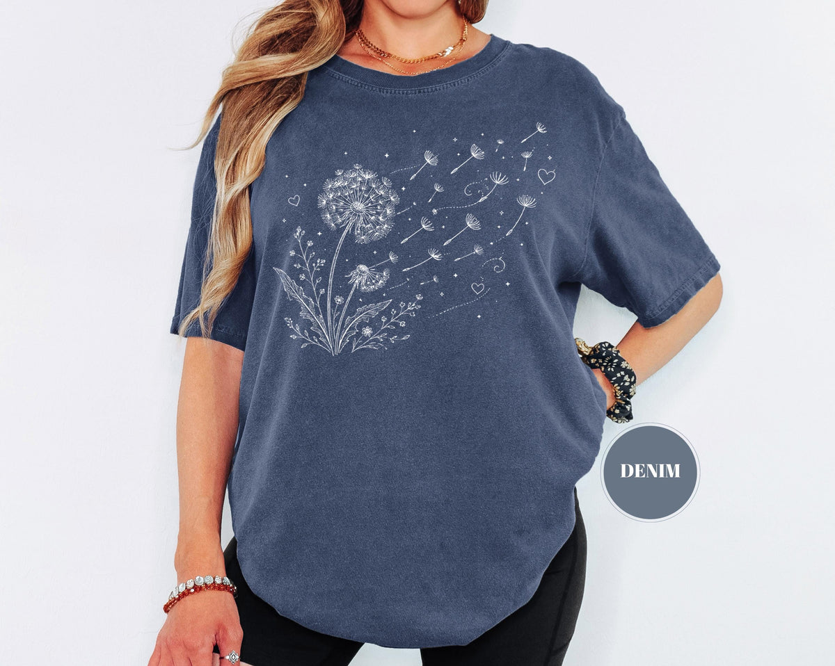 May include: A denim blue t-shirt featuring a white dandelion design with seeds blowing in the wind. The graphic includes hearts and floral elements. The shirt has short sleeves and a round neckline. The word "DENIM" is printed on a circular label.