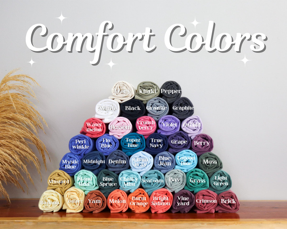 May include: A pyramid of rolled-up t-shirts in various colors, including white, black, and blue. The text "Comfort Colors" is displayed above the shirts. The shirts are arranged on a wooden surface.
