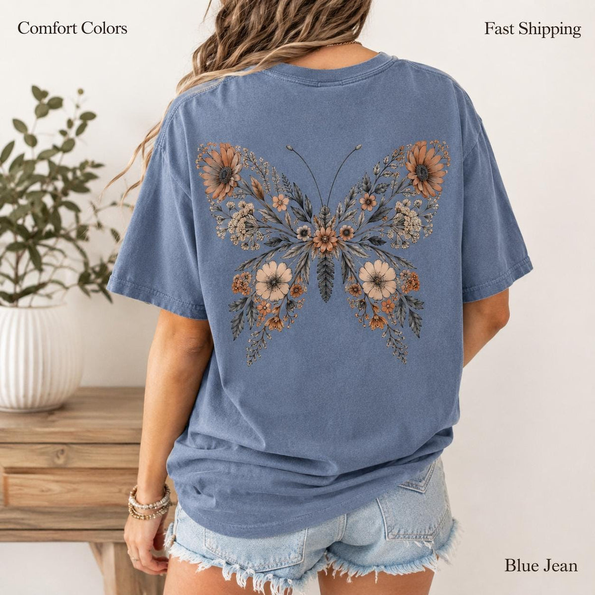 May include: A blue short-sleeve t-shirt featuring a large butterfly graphic on the back. The butterfly is composed of various brown and tan floral illustrations, including sunflowers and small blossoms, with dark stems and leaves.